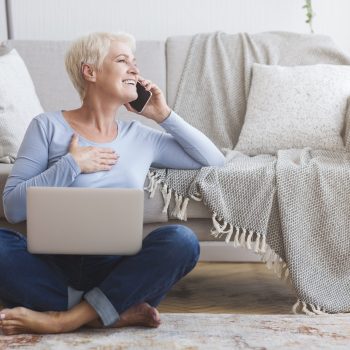 Cheerful elderly business woman talking by phone with clients from home, sitting on floor and using laptop, free space